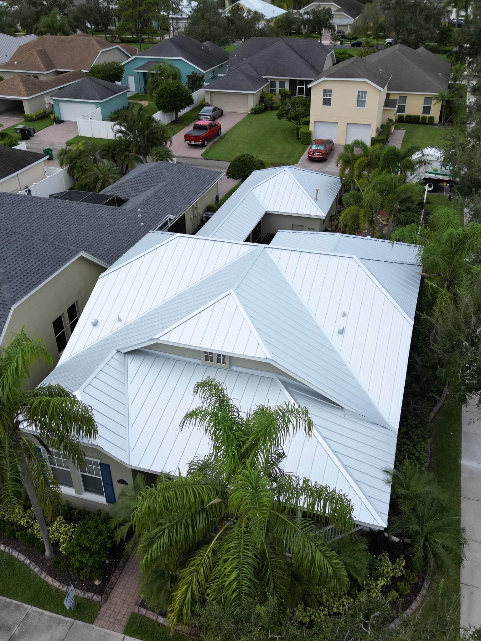 Aerial view of a residential neighborhood with several houses, featuring a white-roofed house surrounded by greenery and parked cars on the driveways.